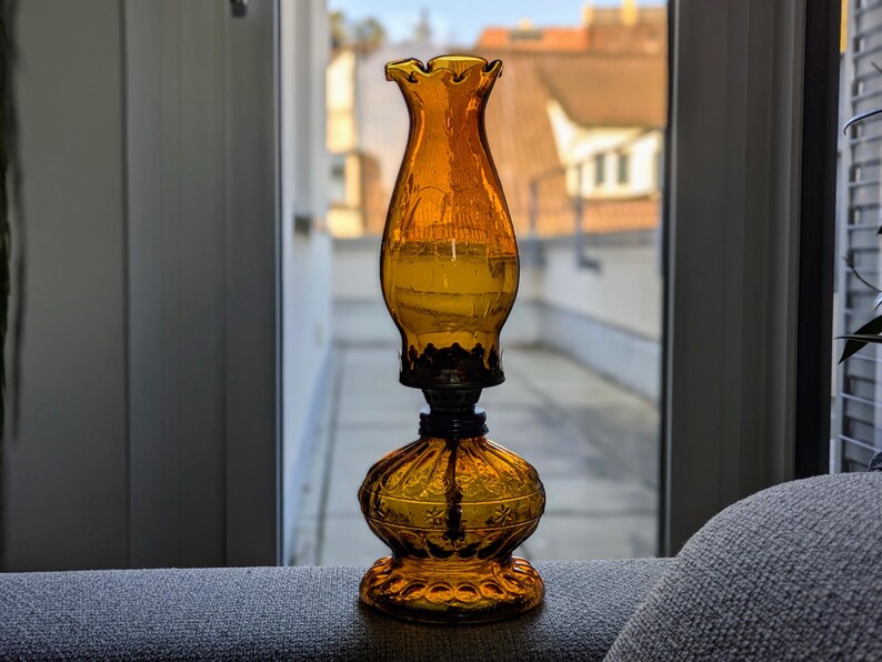 May include: An amber-colored glass oil lamp with a ruffled top and ornate base. The lamp has a dark metal connector and a decorative floral pattern on the base. The lamp is sitting on a gray surface in front of a window.
