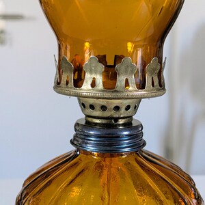 May include: Amber-colored glass oil lamp with a decorative metal crown and a ribbed base. The lamp has a vintage aesthetic, with a brass-colored metal accent around the wick. The lamp is designed to hold oil for illumination.