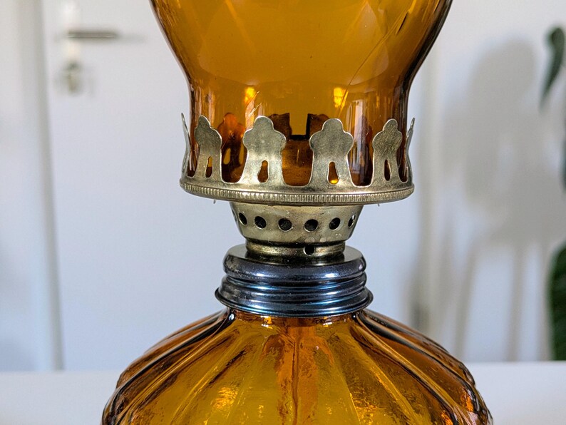 May include: Amber-colored glass oil lamp with a decorative metal crown and a ribbed base. The lamp has a vintage aesthetic, with a brass-colored metal accent around the wick. The lamp is designed to hold oil for illumination.