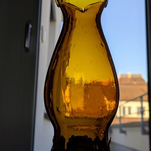 May include: Amber-colored glass lamp shade with a ruffled top and a decorative base. The shade has a classic, vintage design, and the glass appears to be slightly textured. The background shows a window with a view of a building and a clear blue sky.