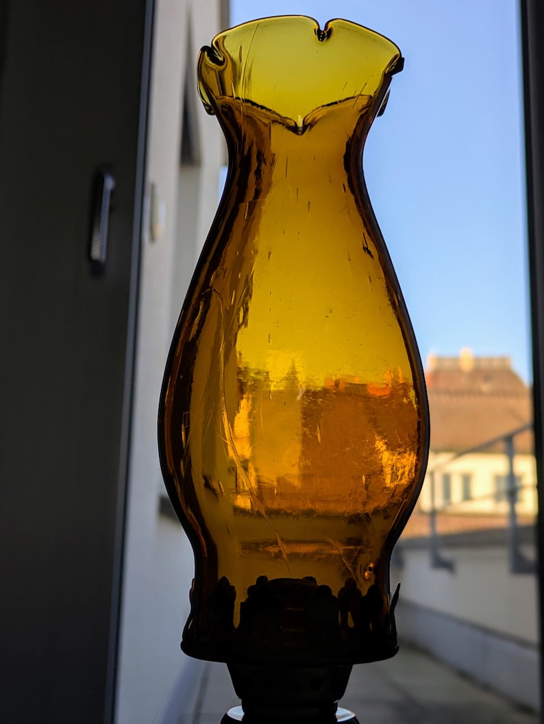 May include: Amber-colored glass lamp shade with a ruffled top and a decorative base. The shade has a classic, vintage design, and the glass appears to be slightly textured. The background shows a window with a view of a building and a clear blue sky.