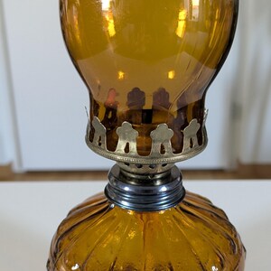 May include: Amber-colored glass oil lamp with a decorative metal base and a ribbed, rounded bottom. The lamp has a bulbous top and a metal collar. The lamp is designed to hold oil and a wick for illumination.