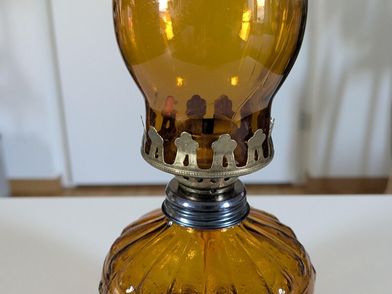 May include: Amber-colored glass oil lamp with a decorative metal base and a ribbed, rounded bottom. The lamp has a bulbous top and a metal collar. The lamp is designed to hold oil and a wick for illumination.