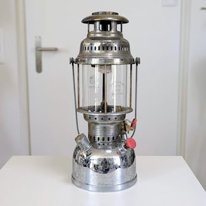 May include: A vintage silver lantern with a clear glass cylinder. The lantern has a red valve and a handle. It is on a white surface. The lantern is designed for outdoor use. The height is approximately 30 cm.