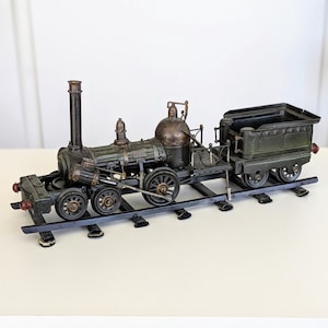 Pode incluir: Uma locomotiva a vapor em miniatura feita de metal em tons de verde escuro e bronze com um tender, posicionada sobre um curto trecho de trilhos de metal preto. A máquina apresenta uma chaminé alta, caldeira arredondada e rodas detalhadas.