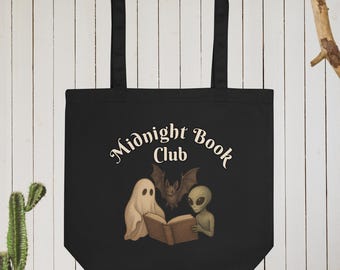 Puede incluir: Bolso tote negro con las palabras "Midnight Book Club" en una fuente caprichosa. El bolso presenta un gráfico de un fantasma, un murciélago y un extraterrestre leyendo un libro. El bolso tiene dos asas negras.