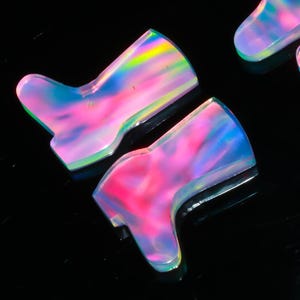 Puede incluir: Dos gemas iridiscentes en forma de bota, con una vibrante exhibición de tonos rosa, azul y amarillo. Las gemas están sobre un fondo negro, resaltando su apariencia colorida y translúcida. Las botas son probablemente para la fabricación de joyas.
