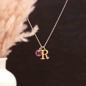 Può includere: Una collana color oro con una catenina sottile e due ciondoli: un pendente circolare con una pietra preziosa rossa scura sfaccettata e un pendente a forma di lettera maiuscola R. I componenti in metallo hanno una finitura lucida.