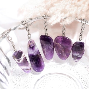 May include: Five polished amethyst stone keychains with silver-toned metal chains and split rings. Each keychain features a unique, irregularly shaped purple gemstone with natural white banding, suspended from a sturdy metal link chain.