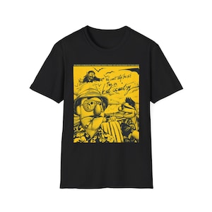 Op de afbeelding: Zwart T-shirt met een gele afbeelding van twee poppenpersonages en de tekst "We can't stop here! This is bat country." De afbeelding bevat een cartoonvleermuis en vogels. Het shirt heeft een ronde hals.