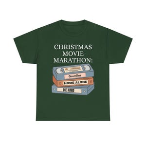 Puede incluir: Camiseta verde oscuro con el texto "CHRISTMAS MOVIE MARATHON:" sobre una pila de cintas VHS. Las cintas están etiquetadas como "Gremlins", "Home Alone" y "Die Hard".