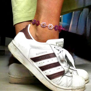 May include: A beaded anklet with flower-shaped beads in shades of purple, pink, and gold. The anklet is worn on an ankle above a white Adidas Superstar sneaker with brown stripes and the word "SUPERSTA" printed on the side.