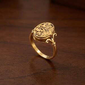 May include: A gold ring featuring an oval-shaped face with an intricate, dark-toned engraved floral pattern. The band connects to the oval head with decorative, curved metal accents that extend from the sides of the ring shank to the top.