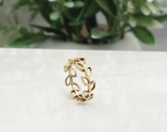 May include: A gold-colored ring featuring a delicate leaf design. The ring's band is adorned with multiple leaf shapes, creating a nature-inspired aesthetic. The ring is set against a white surface with a blurred background of greenery.