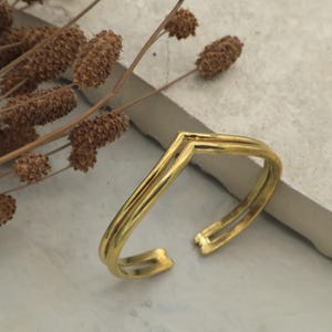 May include: A gold-coloured cuff bracelet with a V-shaped design. The bracelet has a layered appearance and an open design. The background includes dried plants and a neutral-coloured surface.
