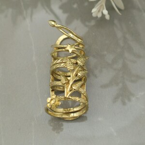 Puede incluir: Un anillo de dedo dorado con múltiples bandas y detalles decorativos florales y de estrella. El anillo presenta una serie de bandas conectadas con un diseño floral y un acento en forma de estrella. El anillo está diseñado para cubrir una parte significativa del dedo.
