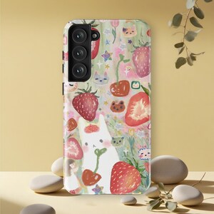 May include: A phone case with a whimsical design featuring a white cat holding a cherry, surrounded by strawberries, cherries, and cat faces. The case has a colorful, pastel background with stars and flowers.