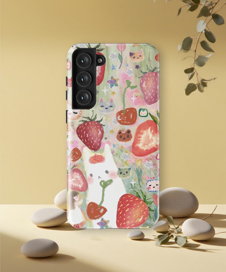 May include: A phone case with a whimsical design featuring a white cat holding a cherry, surrounded by strawberries, cherries, and cat faces. The case has a colorful, pastel background with stars and flowers.