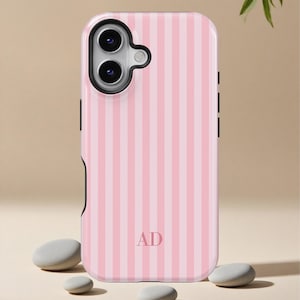 Puede incluir: Una funda de teléfono a rayas rosas y blancas con las iniciales "AD" en el centro. La funda tiene una carcasa de cámara negra y botones laterales negros. La funda está sobre una superficie beige con tres piedras grises pequeñas y lisas.