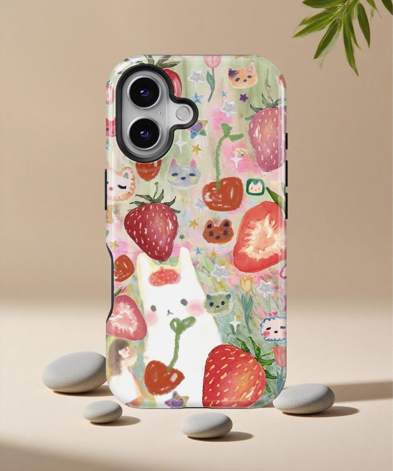 May include: A phone case with a whimsical design featuring strawberries, cats, and other cute illustrations. The case has a white background with a colorful pattern of red strawberries, green leaves, and various cartoon characters. The case is designed to fit a smartphone.