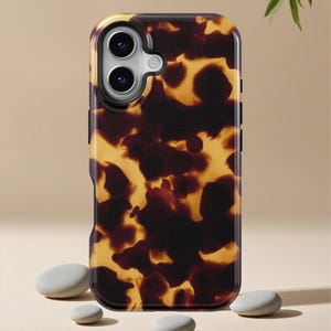May include: A protective phone case with a tortoiseshell pattern in shades of brown and yellow. The case has a camera cutout and a black border. The background is a neutral beige with small gray stones.