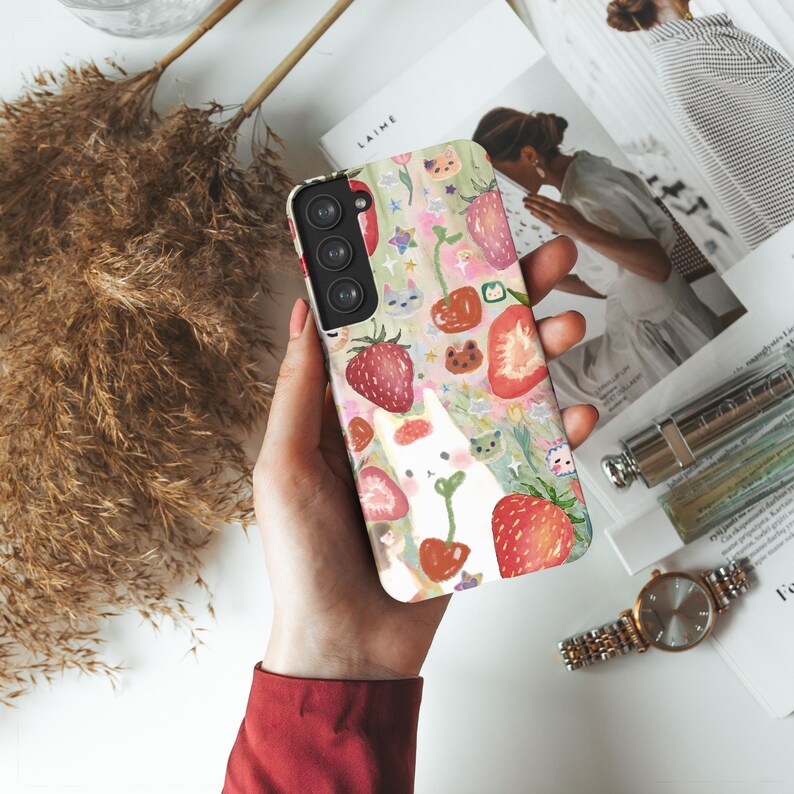 May include: A phone case with a colorful strawberry and cat design. The case features a repeating pattern of red strawberries, white cats, and green foliage on a light pink background. The phone case is held in a person's hand.