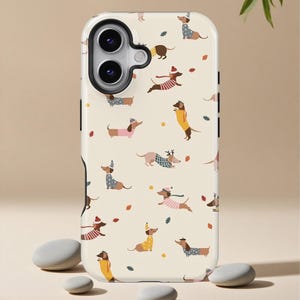 May include: A phone case featuring a repeating pattern of dachshunds wearing various outfits and accessories, such as sweaters and hats, on a cream-colored background. The design includes scattered autumn leaves. The case has a glossy finish and a camera cutout.