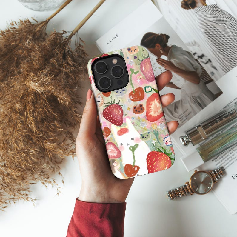 May include: A colorful phone case featuring a whimsical design of strawberries, cherries, and a white cat. The case has a glossy finish and a protective design. The background includes a floral pattern with green leaves and pink flowers.