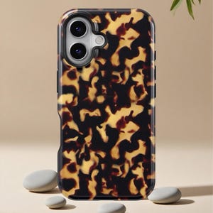May include: A protective phone case with a tortoiseshell pattern in shades of brown and yellow. The case has a black border and a camera cutout. The phone case is shown on a neutral background with small, smooth stones.
