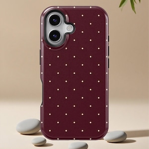 May include: A burgundy phone case with a white polka dot pattern. The case has a black border and a camera cutout. The phone case is shown with several small, smooth, gray stones.