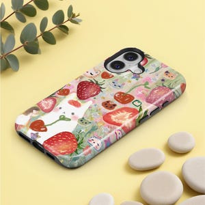 May include: A phone case with a whimsical design featuring strawberries, a white bunny, and other floral elements. The case has a glossy finish and a black border. The overall color scheme is pastel with red, pink, and green accents.