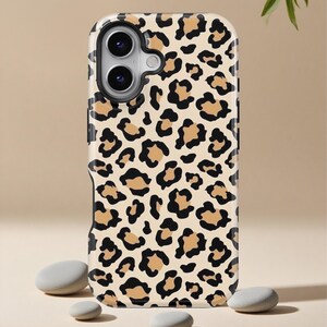 Puede incluir: Una funda de teléfono con un diseño de estampado de leopardo. La funda presenta un fondo color crema con manchas negras y marrón claro. La funda tiene un acabado brillante y está diseñada para adaptarse a un teléfono inteligente. La funda se muestra con tres piedras grises.