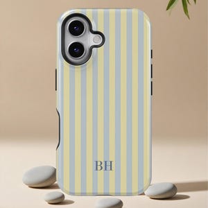 May include: A phone case with a vertical striped pattern in pale yellow and light blue. The case has a black camera cutout and the initials "BH" in navy blue. The case is shown with smooth, gray stones.