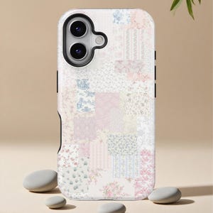 May include: A white phone case with a patchwork design featuring floral patterns in pink, blue, and white. The case has a black border and a camera cutout. Small, smooth grey stones are in the foreground.