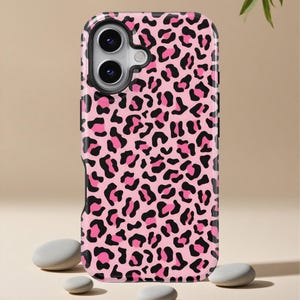 Puede incluir: Funda de teléfono con estampado de leopardo rosa y negro. La funda tiene un borde negro y un diseño de estampado de leopardo rosa. La funda del teléfono se muestra sobre un fondo neutro con piedras pequeñas y lisas.
