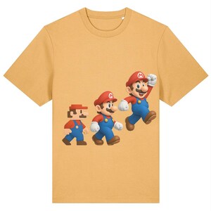 May include: Mustard yellow t-shirt featuring a graphic of the character Mario in three different animated styles. The character is wearing a red hat, blue overalls, and a mustache. The graphic shows Mario in a pixelated form, a 3D form, and a running pose.
