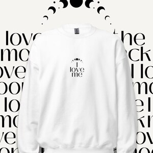 Könnte beinhalten: Weißes Sweatshirt mit dem schwarzen Text "I love me" mittig auf der Brust. Über dem Text befindet sich ein Halbmond-Design. Der Hintergrund zeigt die Worte "love me" in einem sich wiederholenden Muster.
