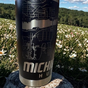 May include: A black stainless steel tumbler with a map of Hancock, Michigan, etched on the side. The tumbler has a silver base and a clear lid. The text "MICHIGAN HUB" is displayed in large, silver letters.