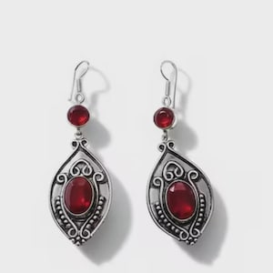 May include: A pair of silver-toned dangle earrings featuring a small round red stone at the top and a larger oval red stone set within an ornate, filigree-style metal frame. The earrings hang from simple silver hooks and have a polished finish.