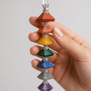 May include: A vertical wand featuring a clear quartz point at the top and bottom, with seven stacked pyramid-shaped stones in red, orange, yellow, green, blue, gray, and purple. Each stone is separated by ornate silver-toned metal filigree caps.
