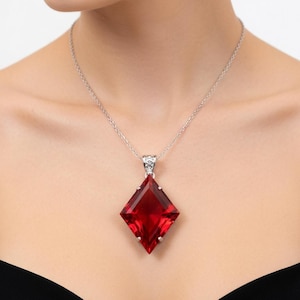 May include: A necklace featuring a large, diamond-shaped, faceted red gemstone pendant suspended from a thin, silver-toned metal chain. The pendant is secured by a small, decorative silver-toned metal bail that connects the stone to the chain.