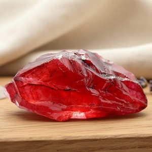 Peut inclure: Une pierre précieuse rouge, facettée et brillante. Le cristal brut est présenté sur une surface en bois, avec un tissu blanc cassé en arrière-plan. La couleur rouge profond de la pierre est mise en valeur par la lumière.