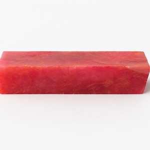 Puede incluir: Una barra rectangular de jabón rojo con aspecto veteado. El jabón tiene una superficie lisa y un borde ligeramente angulado en un lado. El color es un rojo vibrante con toques de naranja.