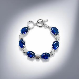 May include: A silver bracelet featuring oval, deep blue gemstones. The bracelet is connected by small, silver flower-shaped links. The clasp is a simple toggle style. The gemstones are evenly spaced around the bracelet.