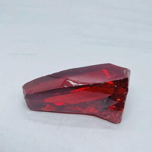 Puede incluir: Un objeto de vidrio rojo oscuro, de forma irregular y facetado. El material translúcido tiene un acabado brillante, lo que sugiere que es un elemento decorativo. El objeto está sobre un fondo blanco, resaltando su color y forma.