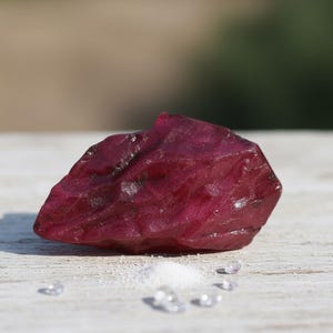 May include: A rough, uncut ruby gemstone with a deep red hue. The ruby is positioned on a white surface, with a small pile of white crystals and several clear, faceted gemstones nearby. The background is blurred, suggesting an outdoor setting.