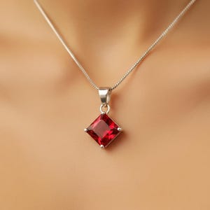 May include: A silver necklace featuring a square, deep red pendant. The pendant is a faceted gemstone set in a silver bezel. The chain is a delicate, thin silver chain.