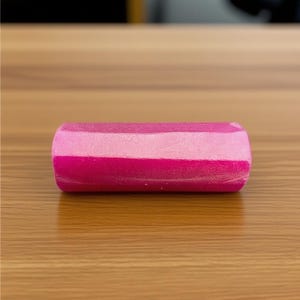 Könnte beinhalten: Ein zylindrisches Objekt mit drei horizontalen Bändern in Rosatönen: hellrosa, mittelrosa und tiefmagenta. Das Objekt liegt auf einer Holzoberfläche.