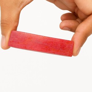May include: A rectangular, red stone, held between two fingers. The stone has a slightly curved shape and a textured surface. The color is a vibrant, solid red with subtle variations in tone. The background is plain white.