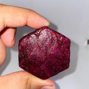Könnte beinhalten: Ein roher, ungeschliffener Rubin-Edelstein. Der Rubin hat eine tiefe, satte rote Farbe mit einer rauen, strukturierten Oberfläche. Der Edelstein wird zwischen zwei Fingern vor einem weißen Hintergrund gehalten. Die Form des Rubins ist unregelmäßig, mit mehreren Facetten.
